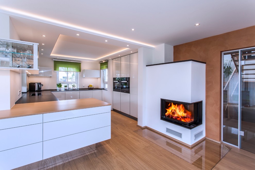 Example of a large trendy u-shaped medium tone wood floor open concept kitchen design in Munich with flat-panel cabinets, a peninsula, a drop-in sink, white cabinets, wood countertops, white backsplash and black appliances