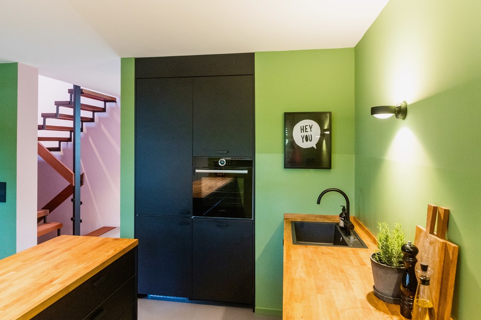 На фото: угловая кухня-гостиная среднего размера в современном стиле с накладной мойкой, черными фасадами, деревянной столешницей, зеленым фартуком, черной техникой, полом из линолеума, островом, серым полом и коричневой столешницей