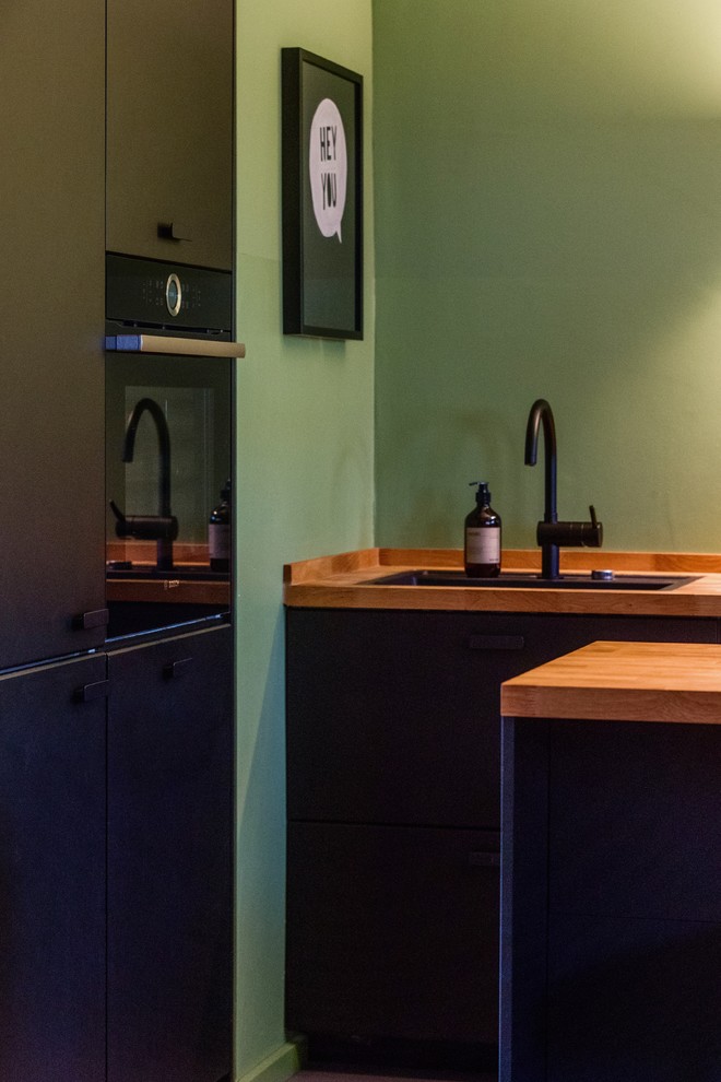 Immagine di una cucina minimal di medie dimensioni con lavello da incasso, ante nere, top in legno, paraspruzzi verde, elettrodomestici neri, pavimento in linoleum, pavimento grigio e top marrone