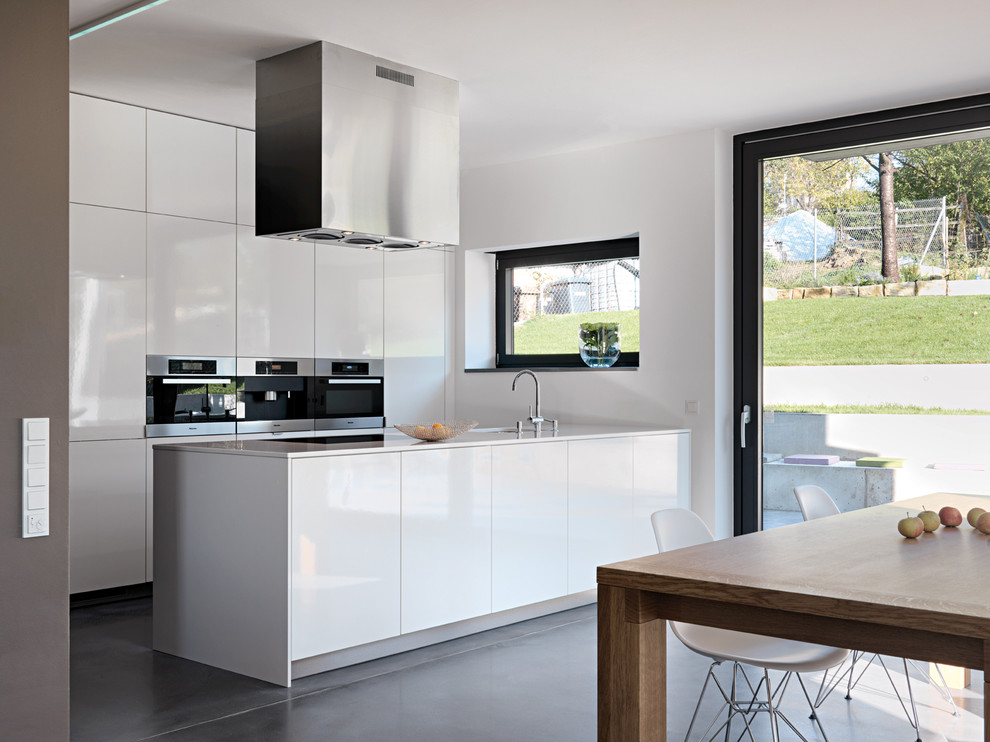 Modern inredning av ett mellanstort kök med öppen planlösning, med rostfria vitvaror, en undermonterad diskho, släta luckor, vita skåp, betonggolv och en halv köksö
