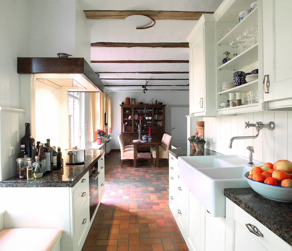 Bild på ett litet lantligt kök, med en rustik diskho, luckor med profilerade fronter, vita skåp, granitbänkskiva, vitt stänkskydd, stänkskydd i keramik, svarta vitvaror och klinkergolv i terrakotta