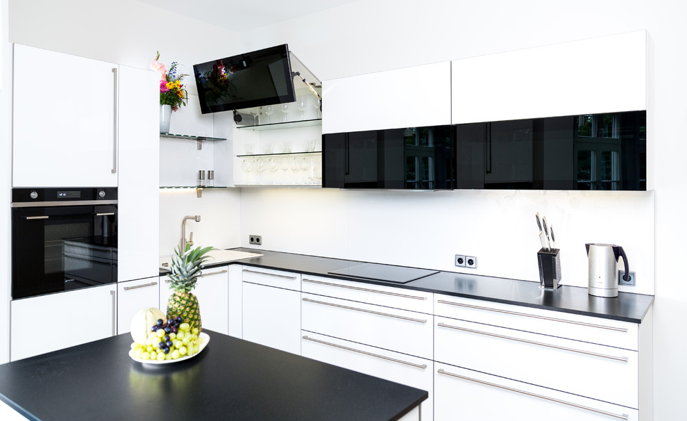 Kleine Moderne Küche in L-Form mit Einbauwaschbecken, flächenbündigen Schrankfronten, weißen Schränken, Küchenrückwand in Weiß, schwarzen Elektrogeräten, Kücheninsel und schwarzer Arbeitsplatte in Sonstige