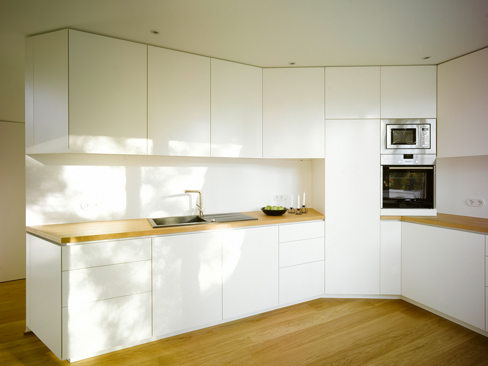 Bild på ett funkis kök med öppen planlösning, med en nedsänkt diskho, släta luckor, vita skåp, träbänkskiva, vitt stänkskydd, rostfria vitvaror och mellanmörkt trägolv