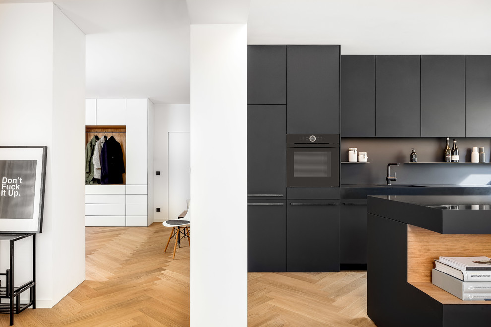 Modelo de cocina lineal moderna con armarios con paneles lisos, puertas de armario negras, salpicadero negro, suelo de madera en tonos medios, una isla y suelo marrón