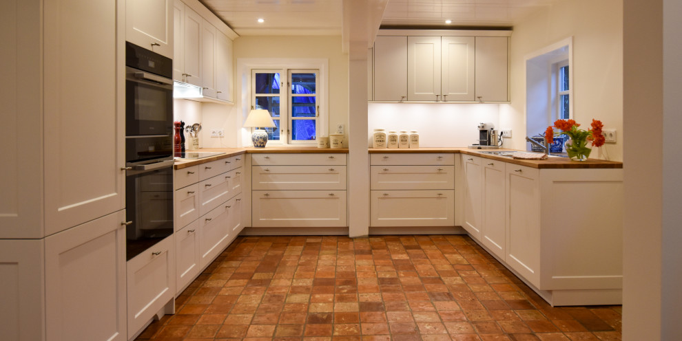 Exempel på ett mellanstort lantligt brun brunt kök, med en nedsänkt diskho, luckor med profilerade fronter, vita skåp, träbänkskiva, svarta vitvaror, klinkergolv i terrakotta och orange golv