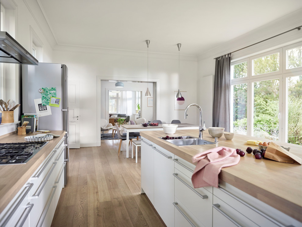 Bild på ett stort nordiskt kök, med en nedsänkt diskho, vita skåp, träbänkskiva, vitt stänkskydd, glaspanel som stänkskydd, rostfria vitvaror, ljust trägolv, en köksö, brunt golv och släta luckor