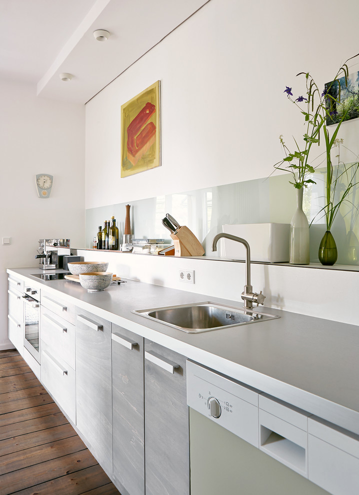 Moderne Küche mit Einbauwaschbecken, flächenbündigen Schrankfronten, grauen Schränken, Küchenrückwand in Weiß, Glasrückwand und braunem Holzboden in Hamburg