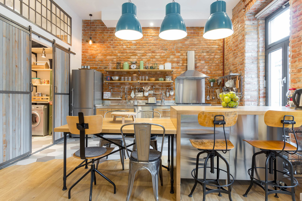 Photo of a medium sized urban galley kitchen in Venice with flat-panel cabinets, medium wood cabinets, stainless steel worktops, brick splashback, stainless steel appliances, light hardwood flooring and a breakfast bar.