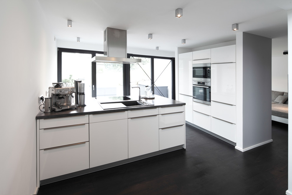 Medium sized contemporary enclosed kitchen in Stuttgart with flat-panel cabinets, white splashback, dark hardwood flooring, a breakfast bar, a built-in sink, white cabinets and stainless steel appliances.