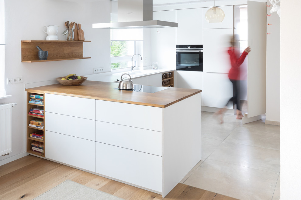 Example of a mid-sized trendy u-shaped limestone floor and beige floor open concept kitchen design in Stuttgart with a drop-in sink, flat-panel cabinets, white cabinets, wood countertops, white backsplash, black appliances, brown countertops and a peninsula