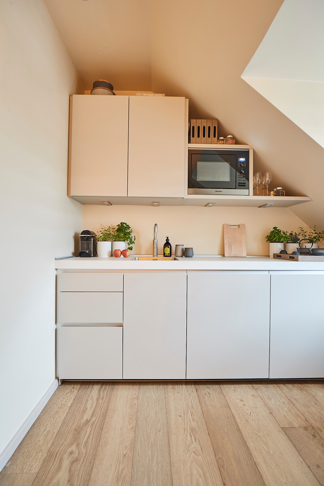 Small contemporary single-wall open plan kitchen in Hamburg with a built-in sink, flat-panel cabinets, beige cabinets, composite countertops, beige splashback, wood splashback, integrated appliances, light hardwood flooring, no island, brown floors and white worktops.