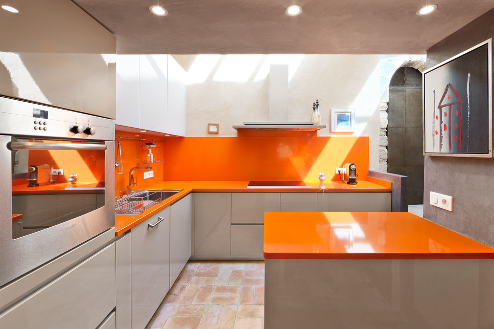 Bild på ett stort funkis orange oranget kök, med en nedsänkt diskho, släta luckor, grå skåp, orange stänkskydd, rostfria vitvaror, en köksö och klinkergolv i terrakotta
