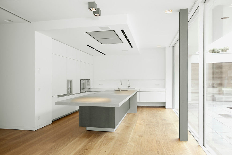 Foto på ett stort funkis l-kök, med en köksö, släta luckor, vita skåp, vitt stänkskydd, ljust trägolv och bänkskiva i betong