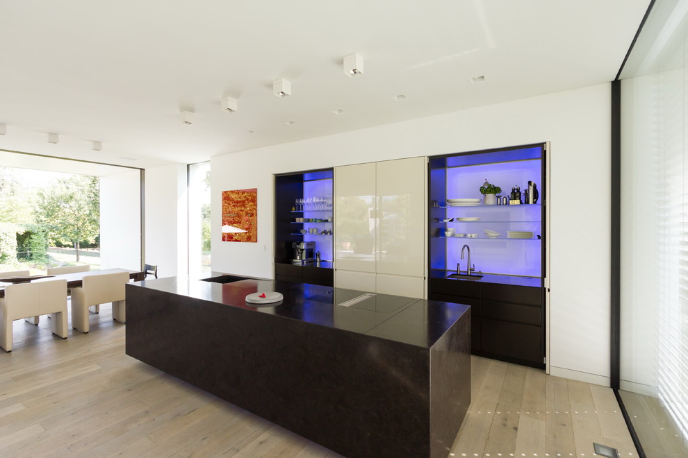 Cette image montre une grande cuisine ouverte parallèle minimaliste avec un évier posé, un placard à porte plane, une crédence bleue, une crédence en feuille de verre, parquet clair, îlot, un sol beige et plan de travail noir.