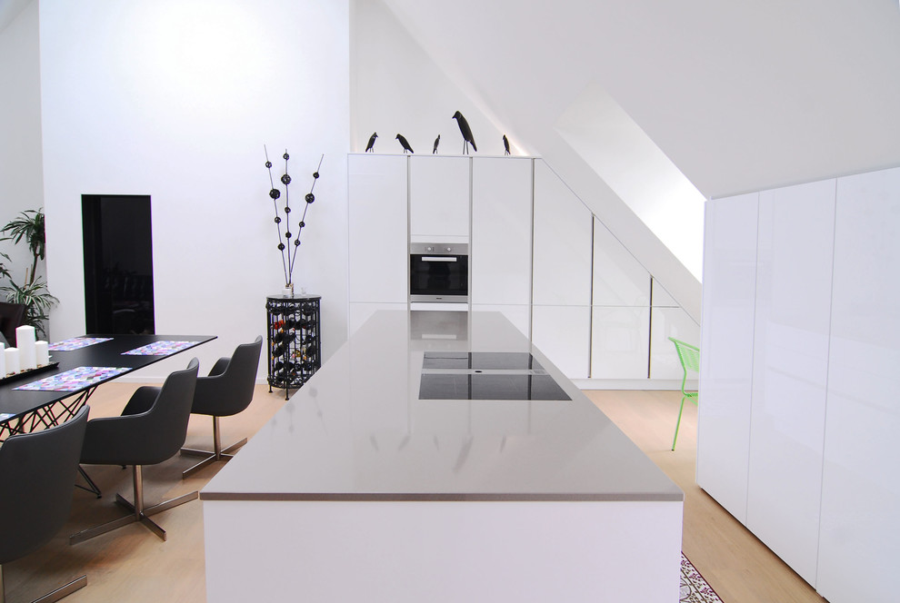 Cette image montre une cuisine parallèle design avec un placard à porte plane, des portes de placard blanches, un plan de travail en quartz modifié, un sol en bois brun, îlot, une crédence blanche et un électroménager noir.