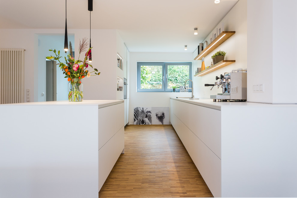 Bild på ett stort funkis vit vitt kök, med en enkel diskho, släta luckor, vita skåp, vitt stänkskydd, glaspanel som stänkskydd, rostfria vitvaror, ljust trägolv, en köksö och brunt golv