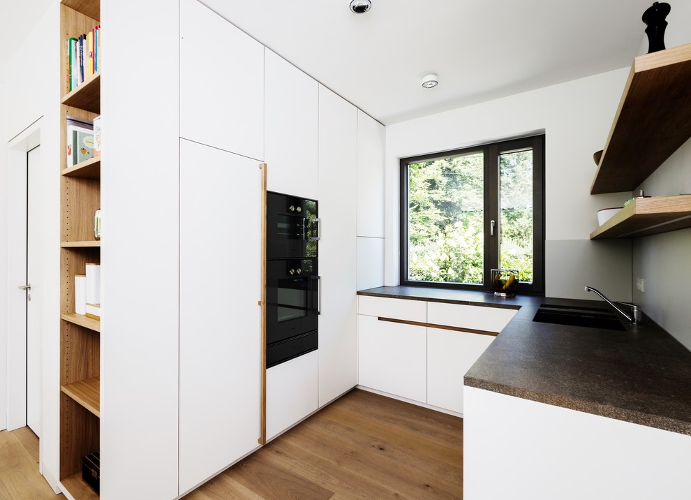 Large contemporary l-shaped open plan kitchen in Munich with a submerged sink, flat-panel cabinets, white cabinets, terrazzo worktops, white splashback, black appliances, medium hardwood flooring, an island and brown floors.