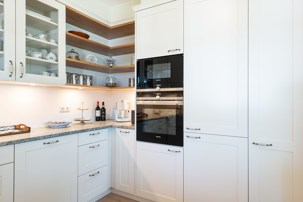 This is an example of a small rural kitchen in Munich with a belfast sink, beaded cabinets, white cabinets, granite worktops, black appliances, medium hardwood flooring, no island, brown floors and grey worktops.
