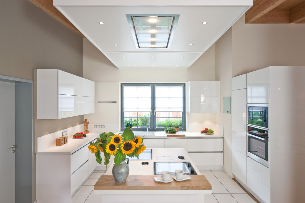 На фото: угловая кухня-гостиная среднего размера в современном стиле с накладной мойкой, плоскими фасадами, белыми фасадами, белым фартуком, техникой из нержавеющей стали и островом