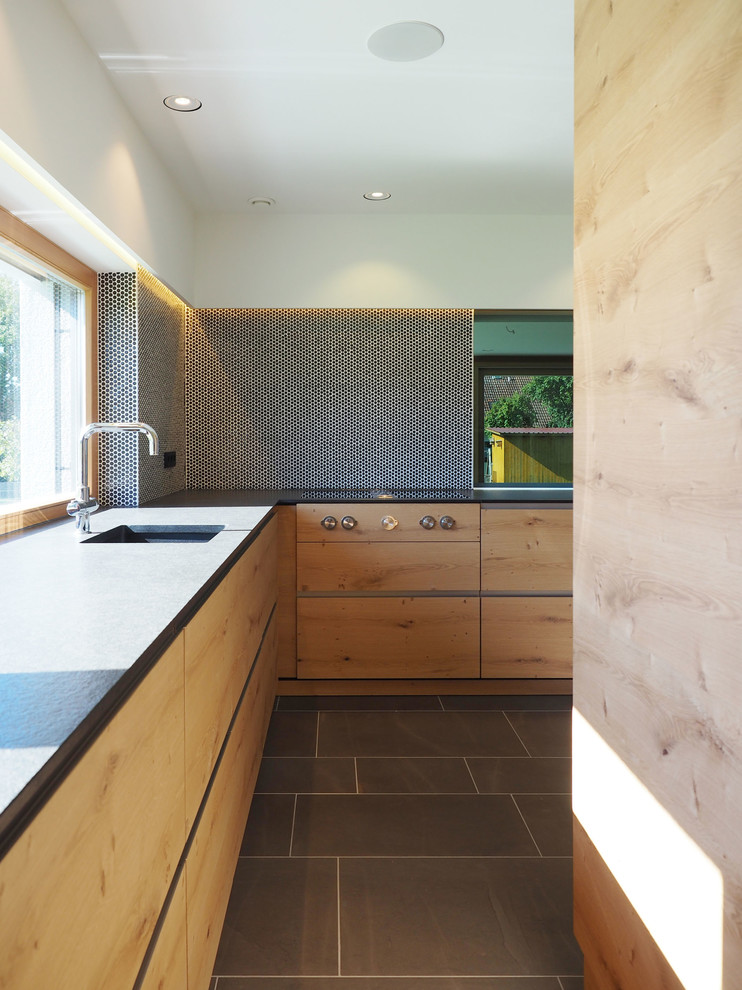 Modern inredning av ett mellanstort, avskilt l-kök, med en integrerad diskho, släta luckor, skåp i ljust trä, stänkskydd i mosaik, granitbänkskiva, svart stänkskydd, rostfria vitvaror och skiffergolv