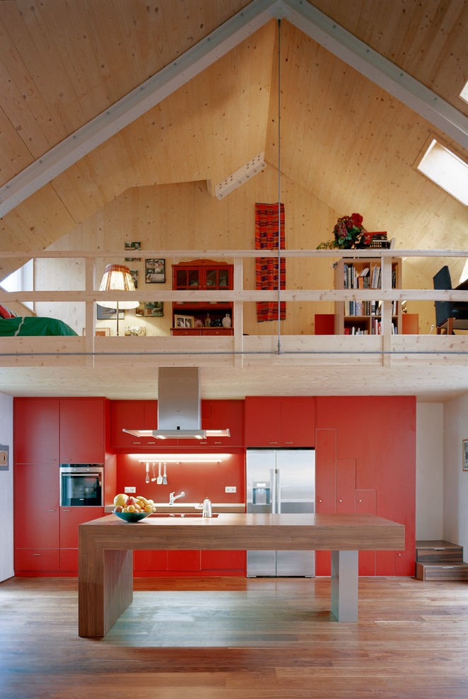 На фото: большая прямая кухня-гостиная в современном стиле с плоскими фасадами, деревянной столешницей, оранжевым фартуком, техникой из нержавеющей стали, паркетным полом среднего тона, островом и красными фасадами