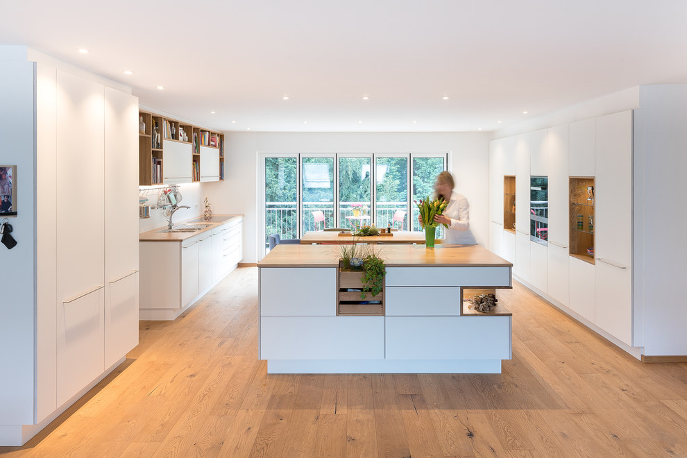 Eat-in kitchen - large contemporary galley medium tone wood floor and brown floor eat-in kitchen idea in Stuttgart with a double-bowl sink, flat-panel cabinets, white cabinets, wood countertops, white backsplash, stainless steel appliances, brown countertops and an island