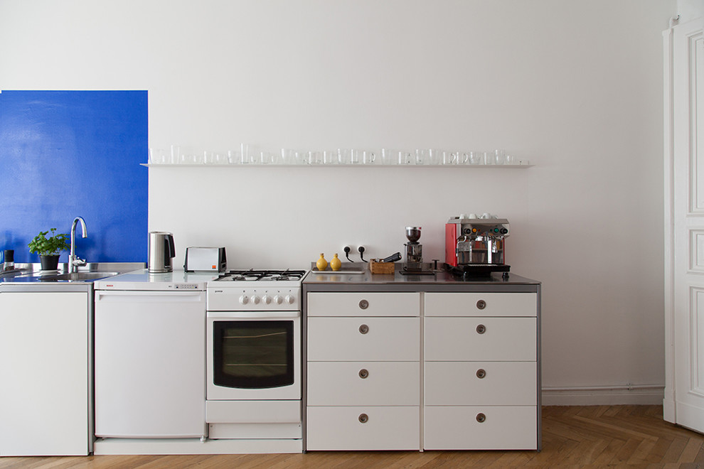 This is an example of a small contemporary single-wall open plan kitchen in Berlin with an integrated sink, flat-panel cabinets, white cabinets, stainless steel worktops, blue splashback, limestone splashback, white appliances, light hardwood flooring, no island and brown floors.