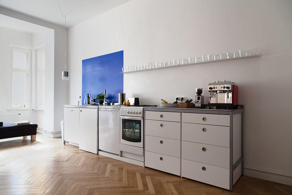 Photo of a small contemporary single-wall open plan kitchen in Berlin with an integrated sink, flat-panel cabinets, white cabinets, stainless steel worktops, blue splashback, limestone splashback, white appliances, light hardwood flooring, no island and brown floors.