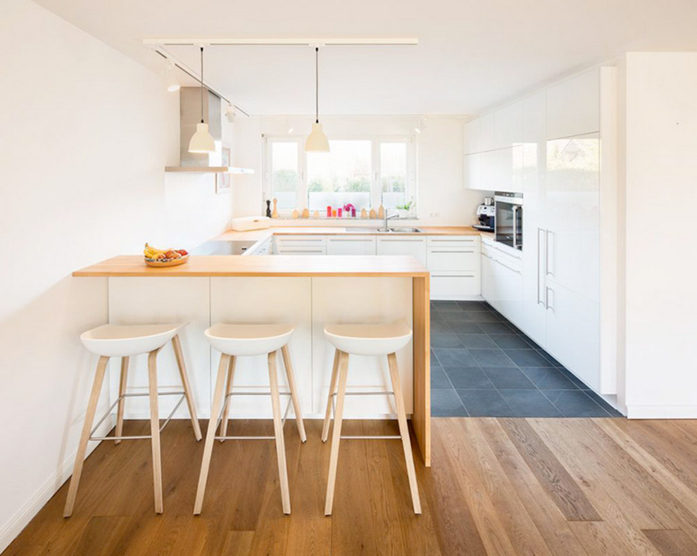 ケルンにある広い北欧スタイルのおしゃれなキッチン (フラットパネル扉のキャビネット、白いキャビネット、木材カウンター、パネルと同色の調理設備、スレートの床、ドロップインシンク) の写真