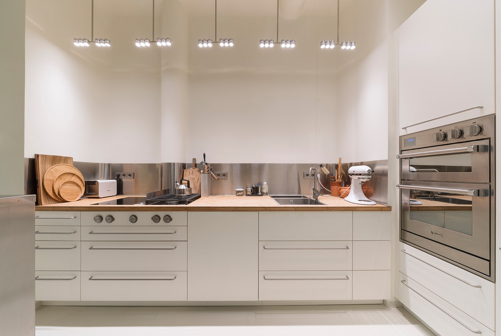 Example of a small trendy l-shaped white floor open concept kitchen design in Hamburg with a double-bowl sink, flat-panel cabinets, white cabinets, wood countertops, metallic backsplash, stainless steel appliances, no island and beige countertops
