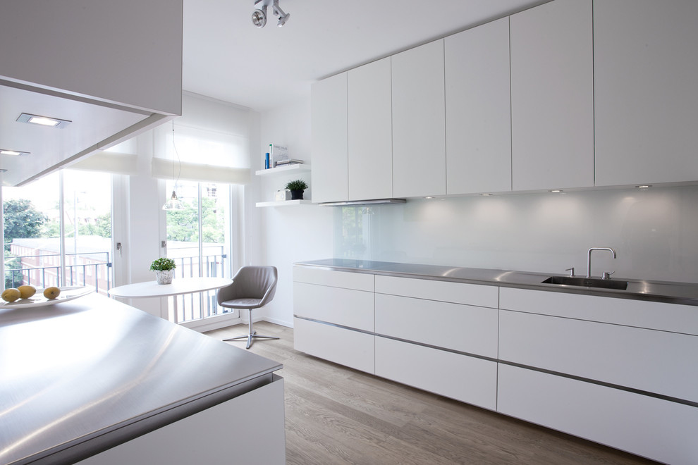 Offene, Mittelgroße Moderne Küche mit integriertem Waschbecken, flächenbündigen Schrankfronten, weißen Schränken, Edelstahl-Arbeitsplatte, Küchenrückwand in Weiß, braunem Holzboden, Kücheninsel und Glasrückwand in München