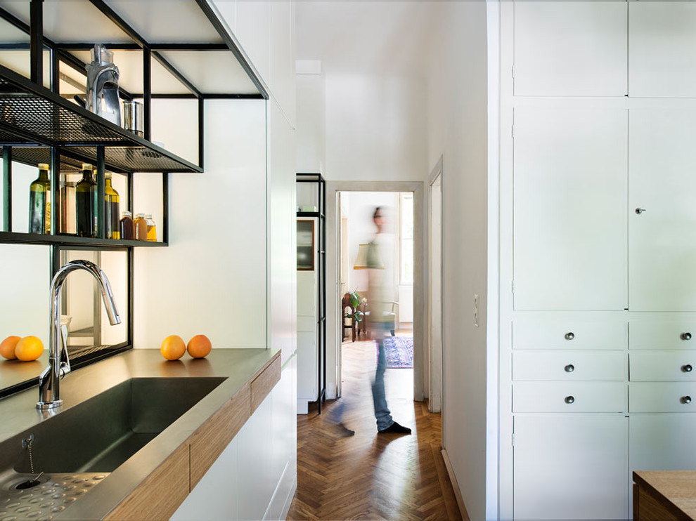 Moderne Küche mit flächenbündigen Schrankfronten, weißen Schränken, Edelstahl-Arbeitsplatte, integriertem Waschbecken und braunem Holzboden in München