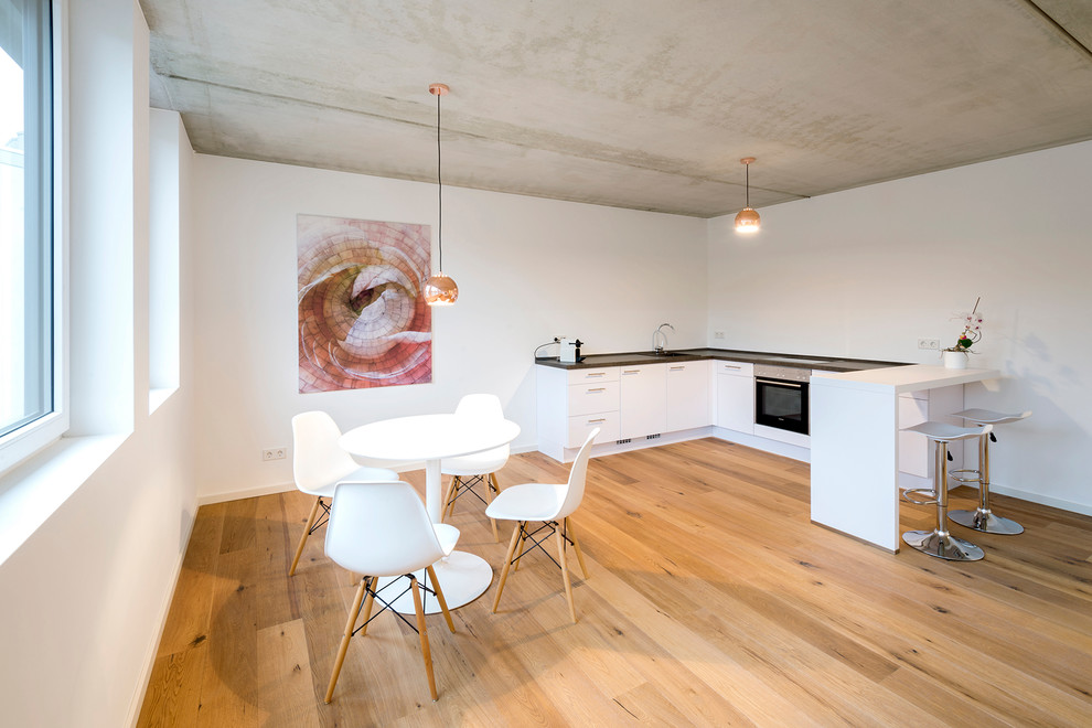 Kleine Moderne Wohnküche ohne Insel in L-Form mit flächenbündigen Schrankfronten, weißen Schränken, Küchenrückwand in Weiß, schwarzen Elektrogeräten und braunem Holzboden in Nürnberg