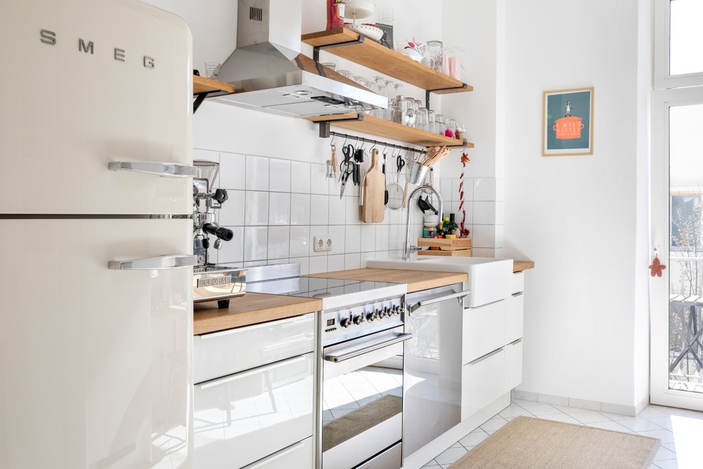 ミュンヘンにある北欧スタイルのおしゃれなI型キッチン (ドロップインシンク、フラットパネル扉のキャビネット、白いキャビネット、木材カウンター、白いキッチンパネル、セラミックタイルのキッチンパネル、白い調理設備、セラミックタイルの床、アイランドなし、白い床、ベージュのキッチンカウンター) の写真