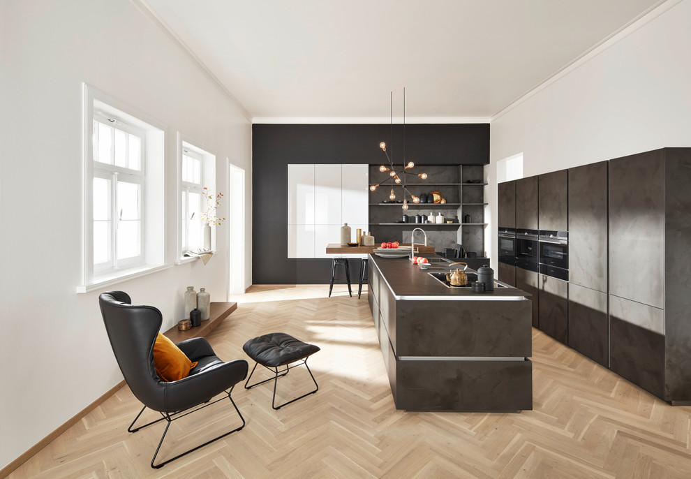 Свежая идея для дизайна: параллельная кухня-гостиная в современном стиле с накладной мойкой, плоскими фасадами, серыми фасадами, серым фартуком, фартуком из сланца, светлым паркетным полом, островом, коричневым полом и черной техникой - отличное фото интерьера