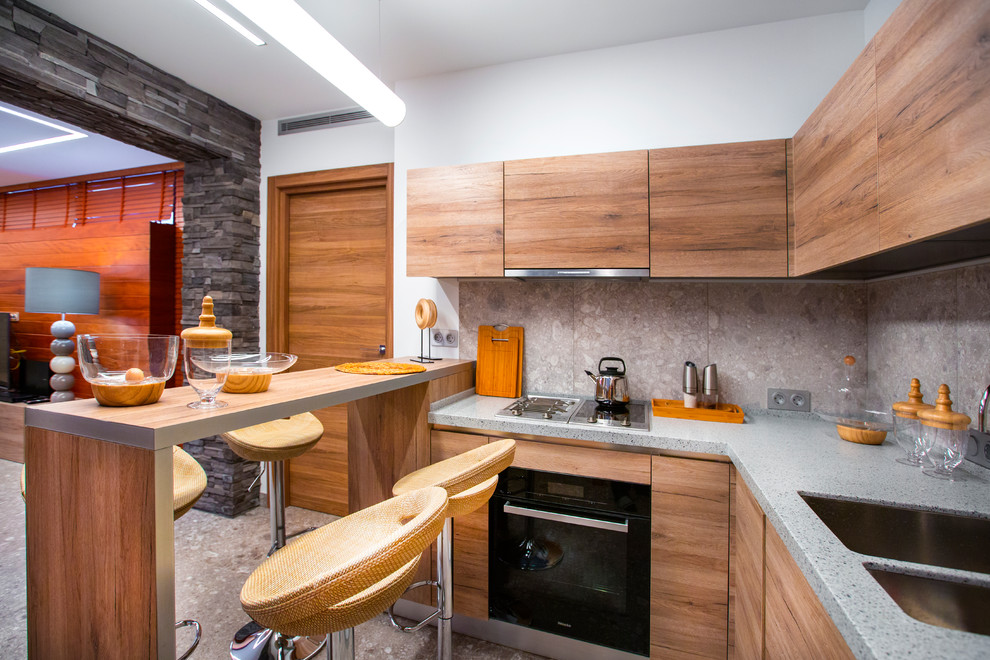Свежая идея для дизайна: п-образная кухня-гостиная в современном стиле с двойной мойкой, плоскими фасадами, фасадами цвета дерева среднего тона, серым фартуком, черной техникой, полуостровом и серым полом - отличное фото интерьера