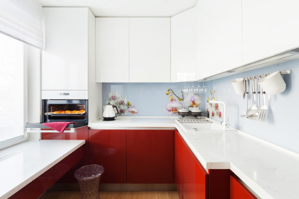 Cette image montre une petite cuisine design en L avec un évier posé, un placard à porte plane, des portes de placard rouges, une crédence multicolore, une crédence en feuille de verre, aucun îlot et un sol marron.