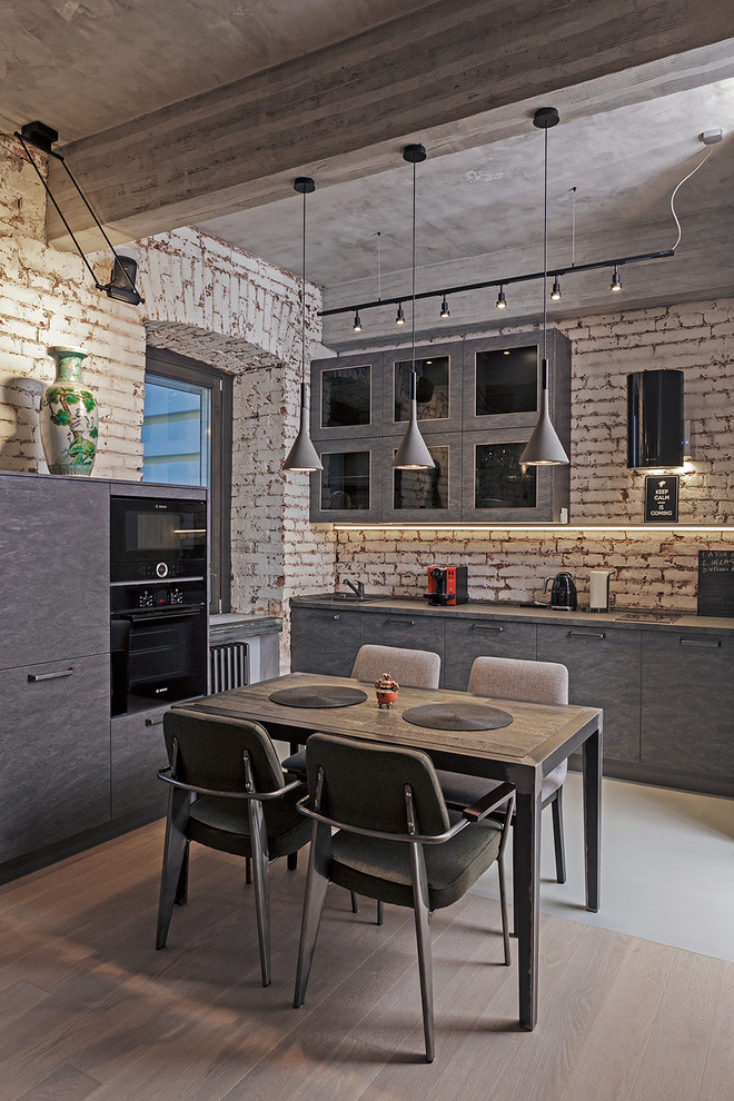 This is an example of a medium sized industrial l-shaped open plan kitchen in Moscow with flat-panel cabinets, grey cabinets, composite countertops, no island, brick splashback, black appliances and light hardwood flooring.