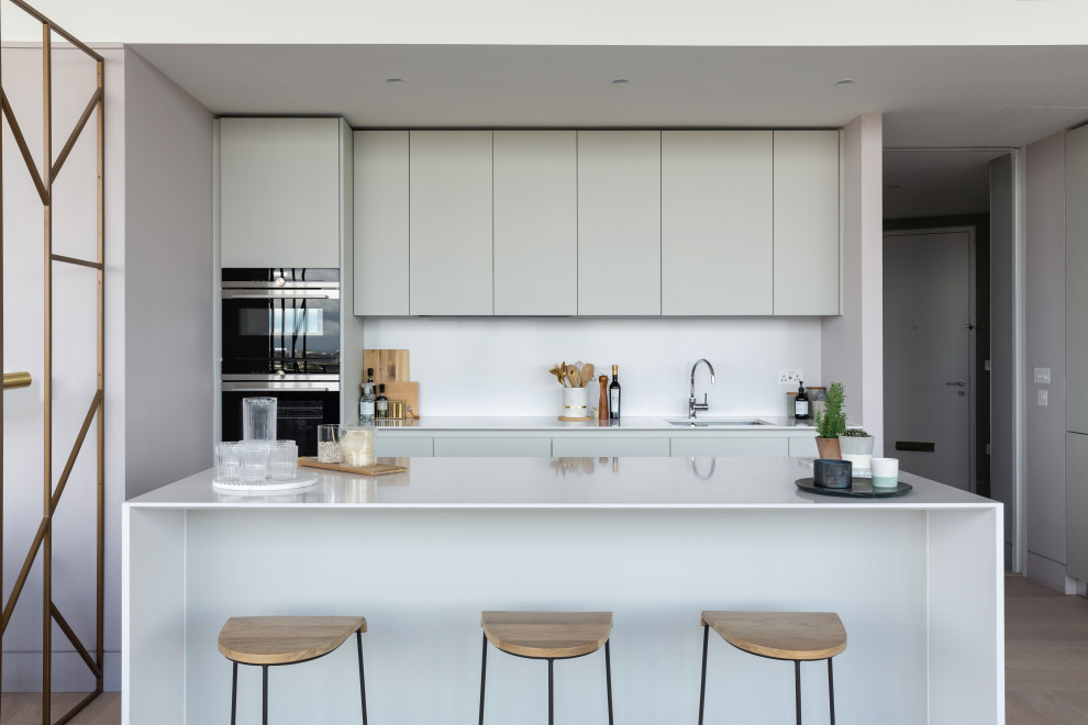Design ideas for a contemporary galley kitchen in London with a submerged sink, flat-panel cabinets, grey cabinets, white splashback, stainless steel appliances, light hardwood flooring, an island, beige floors and white worktops.