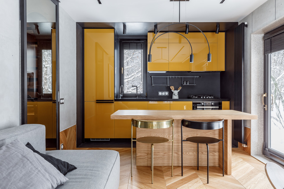 Inspiration pour une petite cuisine ouverte linéaire et bicolore nordique avec un placard à porte plane, des portes de placard jaunes, une crédence noire, un électroménager noir, un sol en bois brun, îlot, un sol marron et plan de travail noir.