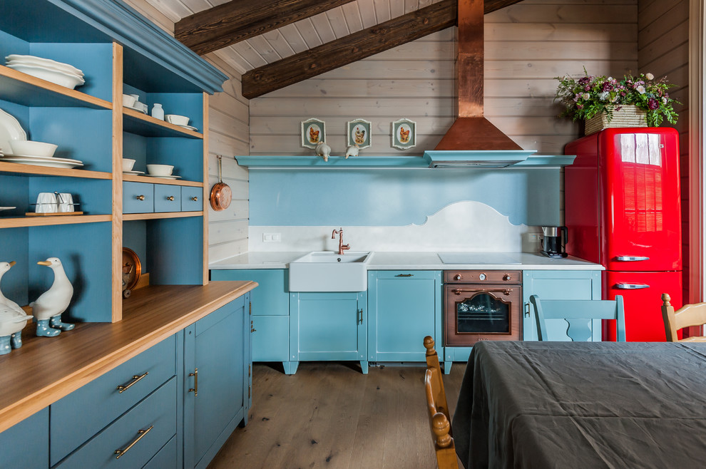 This is an example of a medium sized rural single-wall kitchen/diner in Other with a belfast sink, shaker cabinets, blue cabinets, coloured appliances, brown floors, white splashback, white worktops, composite countertops, stone slab splashback, medium hardwood flooring and exposed beams.