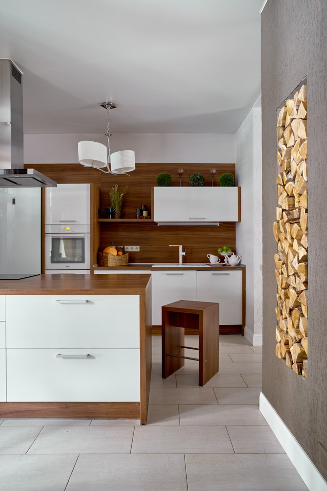 На фото: кухня-гостиная в современном стиле с накладной мойкой, плоскими фасадами, белыми фасадами, коричневым фартуком, белой техникой, островом и серым полом