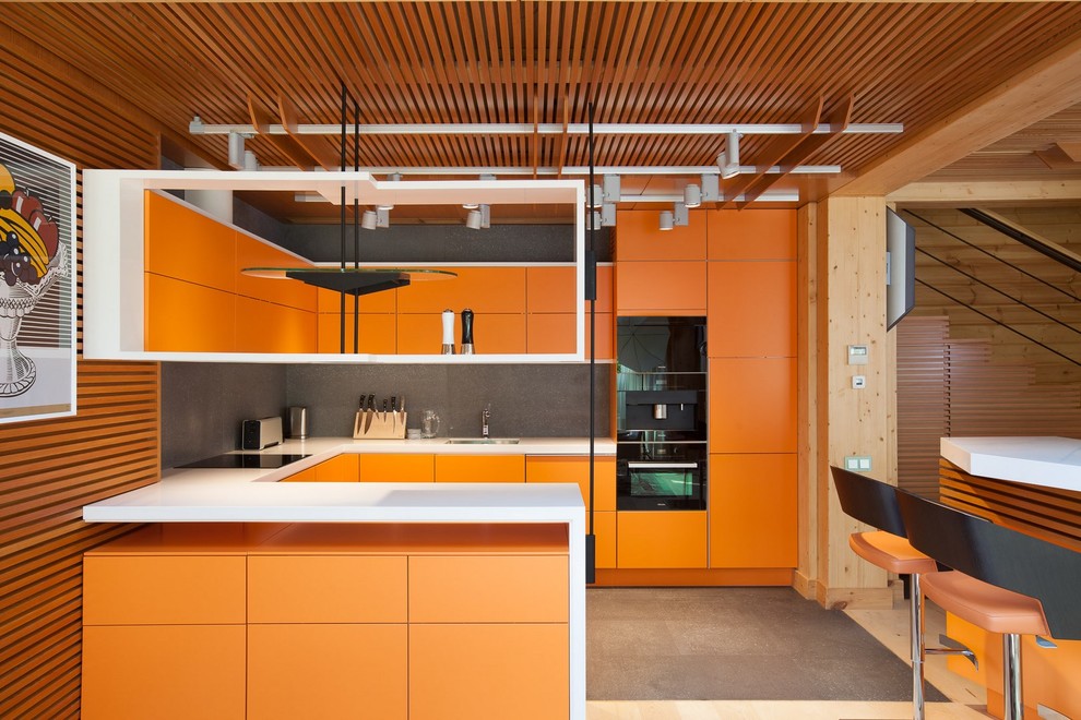 На фото: п-образная кухня-гостиная в современном стиле с врезной мойкой, плоскими фасадами, оранжевыми фасадами, серым фартуком, фартуком из каменной плитки, черной техникой и полуостровом с