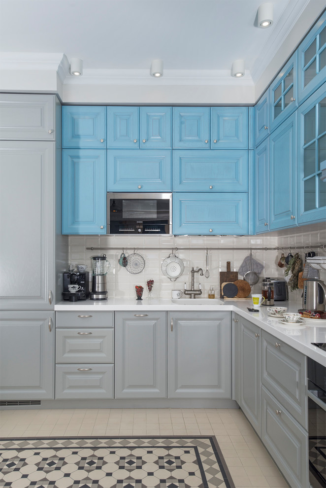 На фото: угловая кухня в стиле неоклассика (современная классика) с фасадами с выступающей филенкой, синими фасадами, белым фартуком, фартуком из плитки кабанчик, черной техникой, бежевым полом и обеденным столом