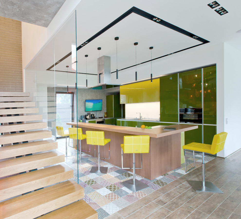 На фото: угловая кухня-гостиная в современном стиле с плоскими фасадами, зелеными фасадами, белым фартуком, черной техникой и островом