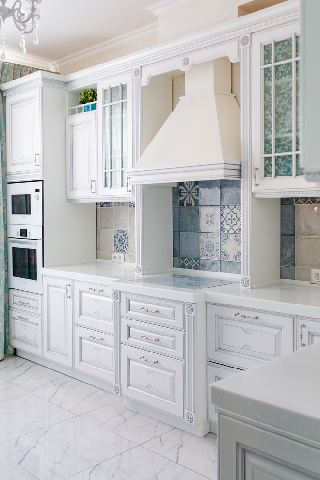 Modelo de cocinas en L clásica con armarios con paneles con relieve, puertas de armario blancas, salpicadero multicolor, electrodomésticos blancos, suelo blanco y encimeras blancas