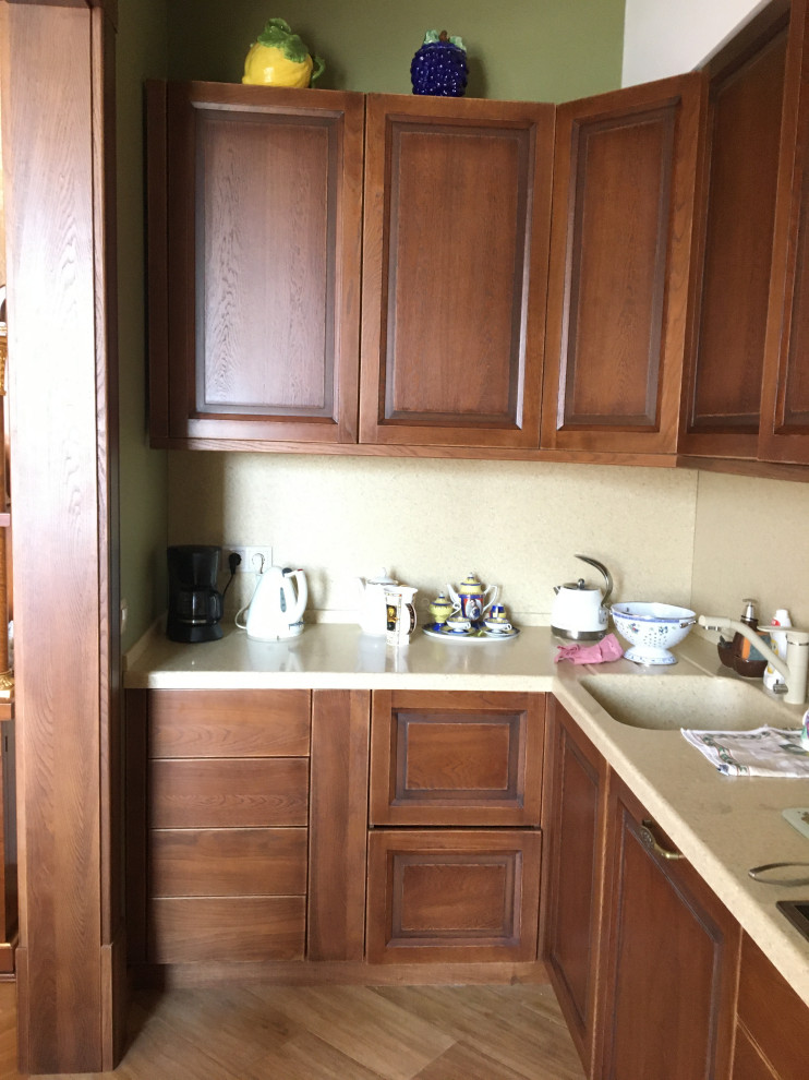 Photo of a medium sized classic l-shaped kitchen/diner in Moscow with raised-panel cabinets, brown cabinets, composite countertops, beige splashback, black appliances, light hardwood flooring, beige floors and beige worktops.