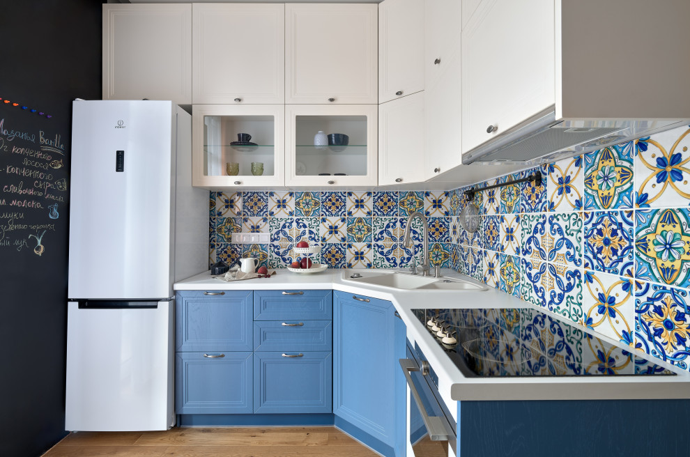 На фото: угловая кухня со шкафом над холодильником в стиле неоклассика (современная классика) с накладной мойкой, фасадами с утопленной филенкой, синими фасадами, белой техникой, паркетным полом среднего тона, коричневым полом, белой столешницей, разноцветным фартуком, двухцветным гарнитуром и мойкой в углу без острова с