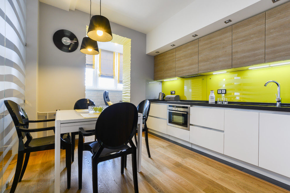 Пример оригинального дизайна: прямая кухня в современном стиле с обеденным столом, плоскими фасадами, белыми фасадами, желтым фартуком, техникой из нержавеющей стали и паркетным полом среднего тона