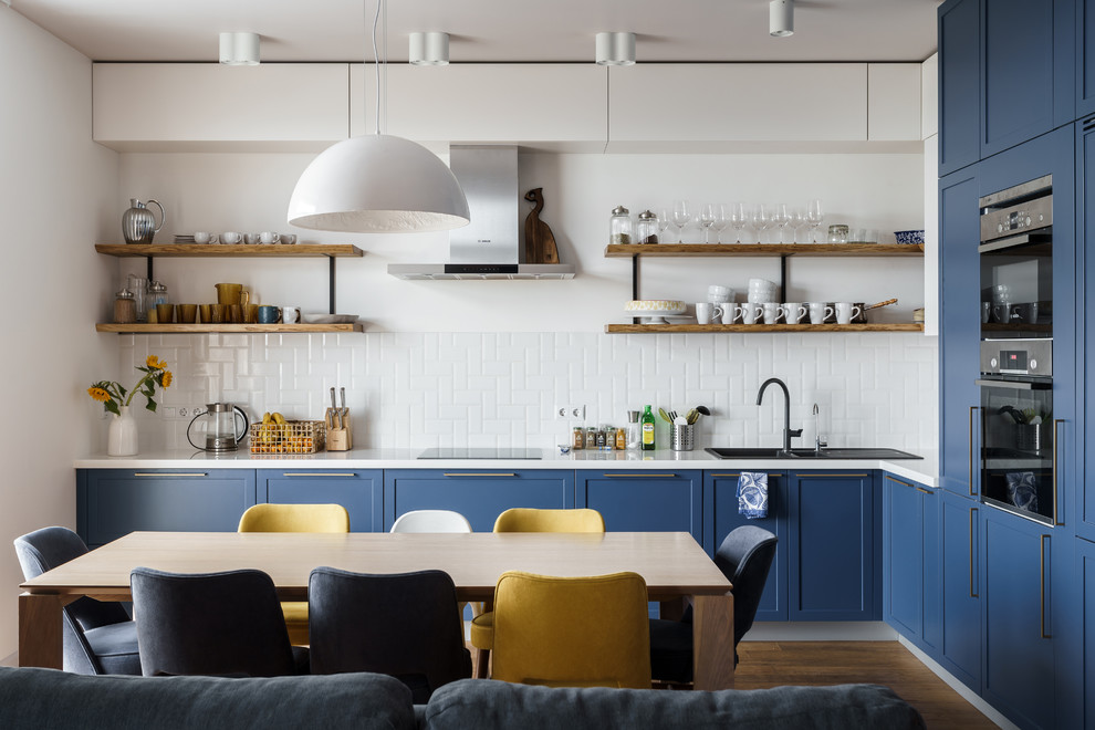 Offene Moderne Küche in L-Form mit blauen Schränken, Küchenrückwand in Weiß, Einbauwaschbecken, Schrankfronten mit vertiefter Füllung, Rückwand aus Metrofliesen, Küchengeräten aus Edelstahl, braunem Boden, weißer Arbeitsplatte und braunem Holzboden in Sonstige